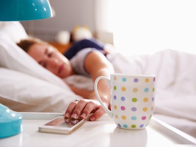 A woman lying in bed with her phone and a cup of coffee on the nightstand. 