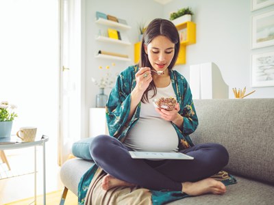 pregnant woman sitting on a couching eating food while looking at an ipad