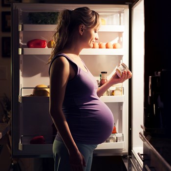 A pregnant woman standing in front of the refrigerator looking at yogurt ingredients.