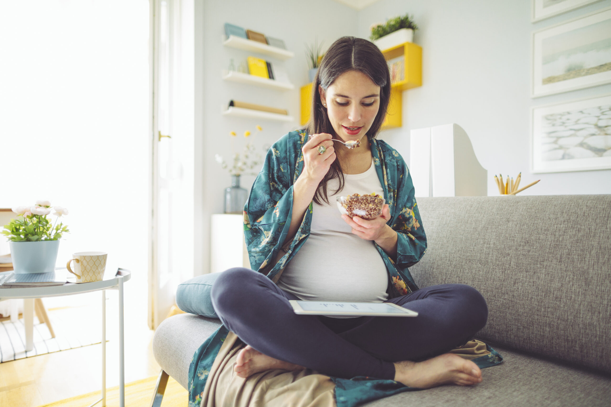 pregnant woman sitting on a couching eating food while looking at an ipad