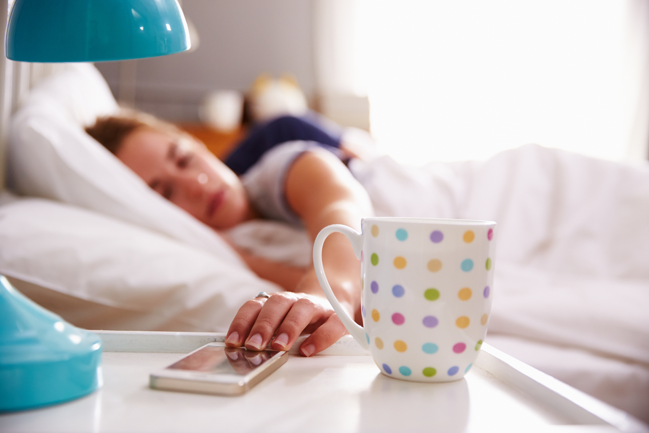 A woman lying in bed with her phone and a cup of coffee on the nightstand.