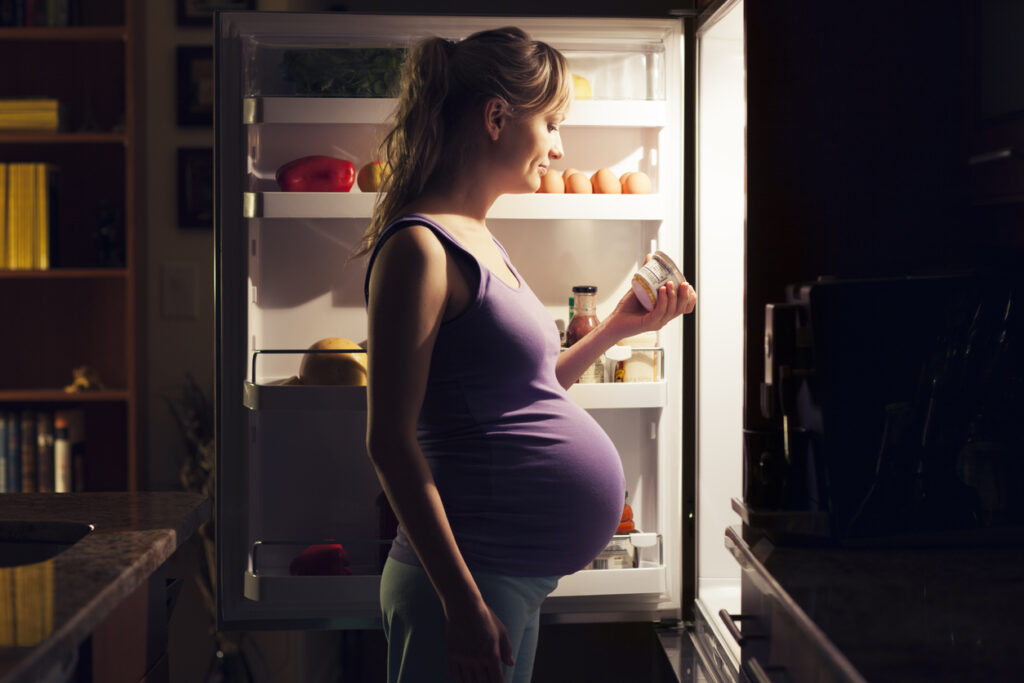A pregnant woman standing in front of the refrigerator looking at yogurt ingredients.