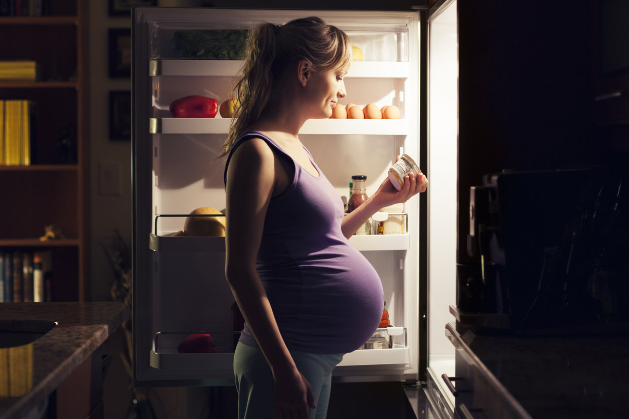 A pregnant woman standing in front of the refrigerator looking at yogurt ingredients.